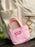 A small pink canvas tote keychain bag with the words 'BUY ME BOOKS' in pink embroidery, resting on a dark wooden surface in front of a cream-colored corduroy backpack.