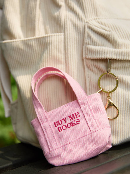 A small pink canvas tote keychain bag with the words 'BUY ME BOOKS' in pink embroidery, resting on a dark wooden surface in front of a cream-colored corduroy backpack.