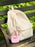 A cream-colored corduroy backpack sits on a dark wooden bench with green foliage blurred in the background. A small pink fabric tiny tote charm with the text 'BUY ME BOOKS' hangs from the side pocket of the backpack.