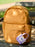 A small lavender and off-white canvas key pouch with a heart-shaped patch reading 'ENEMIES TO LOVERS' hangs from a metal ring on a mustard yellow backpack, resting on a dark bench.
