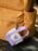 A small lavender and off-white canvas key pouch with a heart-shaped patch reading 'ENEMIES TO LOVERS' hangs from a metal ring on a mustard yellow backpack, resting on a dark bench.