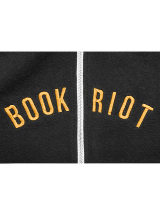 Close-up of a black zip-up garment with the words 'BOOK RIOT' embroidered in yellow across both sides of a white zipper.