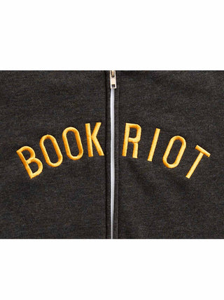 Close-up of a dark charcoal zip-up hoodie with 'BOOK RIOT' embroidered in yellow across the chest, centered on a white background.